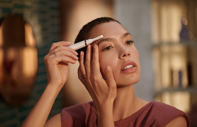: Žena s tmav&yacute;mi vlasy a hněd&yacute;ma očima použ&iacute;v&aacute; př&iacute;stroj Philips k &uacute;pravě chloupků oboč&iacute;.